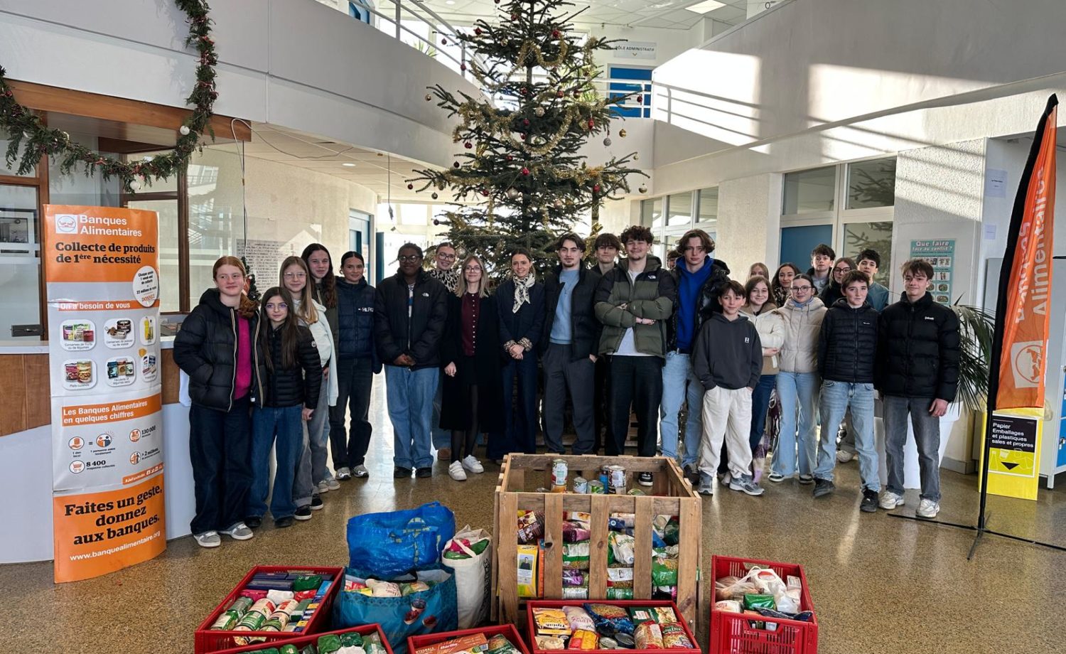 Elèves et adultes du collège souriants et enthousiastes, entourant les boîtes de dons remplies de denrées alimentaires et de produits d’hygiène, symbolisant leur engagement dans la collecte de Noël pour la Banque Alimentaire.